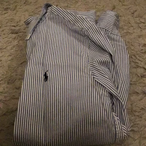 Randig skjorta från Ralph Lauren - Snygg blå- och vitrandig skjorta från Ralph Lauren med klassisk krage och knappar framtill. Den har en broderad logotyp på bröstet.
