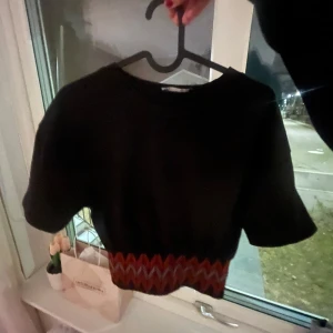 Svart kortärmad topp med mönster - Säljer en svart kortärmad topp med ett färgglatt sicksackmönster i midjan. Perfekt för en trendig look. Passar bra till både jeans, shorts och kjol🥰🥰Säljer för bra pris! Märke: Zara💕💕