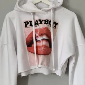 Vit croppad hoodie med Playboy tryck  - Säljer en vit croppad hoodie från Playboy med ett stort tryck av röda läppar på framsidan. Hoodien har en justerbar dragsko i huvan och ribbade muddar vid ärmsluten. Perfekt för en avslappnad stil.