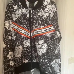  Adidas kofta  - Säljer en snygg svartvit blommig jacka från Adidas med färgglada ränder och logotyp på framsidan. Storlek 164 (13-14år) men passar lika väl xs/ s. Sparsamt använd så i mycket fint skick! 