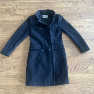 Rock jacka  - Elegant svart kappa med dubbelknäppning och långa ärmar. Tillverkad i en ullblandning med polyesterfoder. Perfekt för kyligare dagar med stilren design och knappar vid ärmsluten. Har ni frågor är det bara att ställa de!!