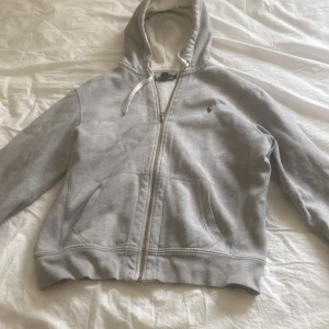 Grå hoodie från Polo Ralph Lauren - Säljer en klassisk grå hoodie från Polo Ralph Lauren med dragkedja och huva. Tröjan har en liten broderad logotyp på bröstet och är tillverkad i en mjuk bomullsblandning. Perfekt för en avslappnad stil.