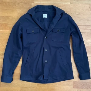 John Henric overshirt - Snygg mörkblå overshirt från John Henric med knappar framtill och två bröstfickor med lock. Jackan har en klassisk krage och långa ärmar med knappar vid ärmsluten. Perfekt för en stilren look.