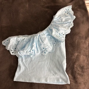 Ljusblå offshoulder topp med volang - Säljer en söt ljusblå offshoulder topp med volangdetaljer. Toppen har ett broderat mönster på volangen och är ribbad för en skön passform. Perfekt för sommarens varma dagar! 💙