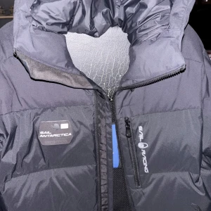 Svart dunjacka från Sail Racing - Säljer en svart dunjacka från Sail Racing med hög krage och dragkedja. Jackan har en stilren design med logotypdetaljer på bröstet och ärmen. Perfekt för kalla dagar.