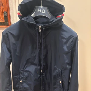 Mörkblå vindjacka från Moncler - Säljer en stilren mörkblå vindjacka från Moncler med huva och dragkedja. Jackan har långa ärmar och två fickor framtill med knappdetaljer. säljer den eftersom att den är för liten för mig. Ingen diskutering om priset