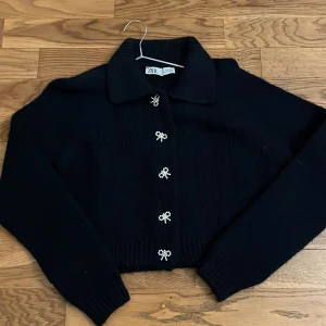 Marinblå  kofta från Zara med pärldetaljer - Snygg marinblå kofta från Zara med unika pärldetaljer i form av rosetter längs knäppningen. Koftan har en klassisk krage och långa ärmar, perfekt för en stilren look. Passar bra till både vardag och speciella tillfällen.
