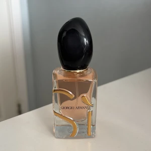 Giorgio Armani Sì Parfym - Säljer en helt ny parfym. Elegant parfymflaska från Giorgio Armani med svart lock och genomskinlig glasflaska. Flaskan har en stilren design med guldiga detaljer och innehåller en ljus doftvätska. Perfekt för den som älskar lyxiga och sofistikerade dofter.
