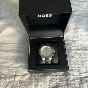 Silverfärgad klocka från BOSS - Snygg och stilren silverfärgad klocka från BOSS. Klockan har en rund urtavla med tre mindre visare och ett robust armband i metall. Perfekt för den som vill ha en elegant accessoar.