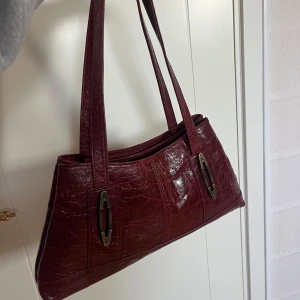 Vinröd axelväska  - Vinröd axelväska i skinn med krokodilpräglat mönster. Köpt vintage