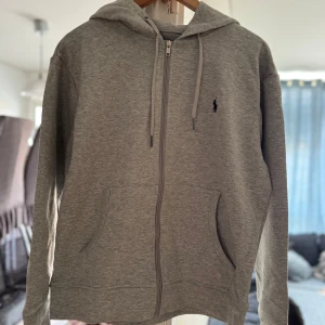 Grå hoodie från Polo Ralph Lauren - Snygg grå hoodie från Polo Ralph Lauren med dragkedja framtill och klassisk logga på bröstet. Tröjan har huva med snörning, två fickor och är tillverkad i mjuk bomullsmix. Perfekt för en chill och stilren look.