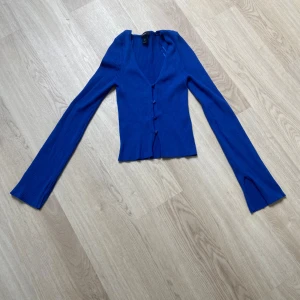 Kornblå ribbad kofta från Lindex - Kornblå ribbad kofta från Lindex med v-ringning och långa ärmar. Modellen har knappar framtill och coola slitsar vid ärmsluten. Sitter tajt och är perfekt för lager-på-lager. Superstretchigt material som känns skönt mot huden. Har bara använt den 1 gång så den är som ny. 