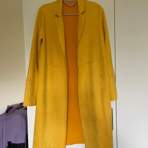 Gul kappa från Zara i mockaimitation - Snygg lång gul kappa från Zara i mockaimitation. Rak modell med klassisk krage och långa ärmar. Perfekt statement-plagg för dig som vill sticka ut med färg. Enkel och stilren design utan knappar eller dragkedja.Oanvänd 