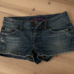 Blå jeansshorts från Zippylugg - Snygga blå jeansshorts från Zippyziggy med låg midja. De har slitna detaljer, knapp och dragkedja framtill samt bakfickor med knapp. Perfekta för varma dagar och en chill stil. En av knapparna saknas på bakfickan.  Midjemått: 39cm. Längd: 23cm