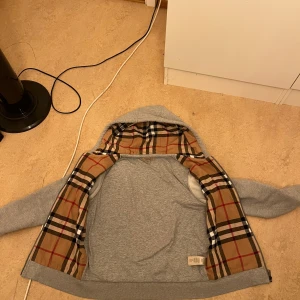 Grå hoodie från Burberry - Snygg grå hoodie från Burberry med klassiskt rutigt foder i beige, svart, vitt och rött. Perfekt för dig som vill ha en stilren look med en touch av lyxigt mönster på insidan. Priset går att diskuteras vid snabb affär.