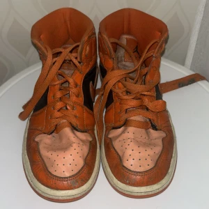 Nike Air Jordan 1 orange/svart - Säljer ett par Nike Air Jordan 1 i orange och svart skinn med klassisk vinge-logga på sidan. Skorna har snörning, perforerad tå och orange yttersula. Om man tvättar dom så är dom nästan som ny😊