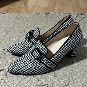 Svartvita rutig pumps med klack - Snygga pumps med spetsig tå och låg klack i svartvitt rutigt mönster. Skorna har en dekorativ rem med spänne över foten och är fodrade i ljusbeige. Perfekta för dig som gillar att sticka ut med klassisk stil och coola detaljer.