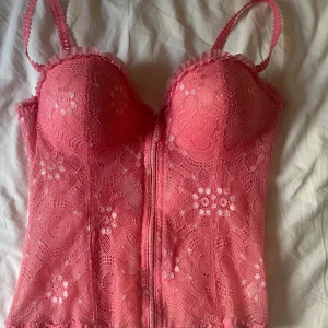 Rosa spetskorsett med rosett och dragkedja - Supersöt rosa korall korsett med blommig spets, justerbara axelband och en snygg dragkedja framtill. Baksidan har en dekorativ rosett och ett litet smycke. Festtopp, nästan aldrig använd. Passar storlek S-M. Push up, passar B-C kupa. KÖP BARA VIS POSTNORD!!!