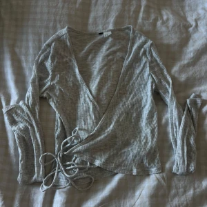 Grå omlottopp med knyt från H&M - Supersnygg grå ribbad omlottopp från H&M (lappen är dock bortklippd pga kli) med långa ärmar och knyt i midjan. Toppens tunna, mjuka bomullsmaterial och diskreta ränder gör den perfekt för lager-på-lager eller som den är. V-ringad och sitter riktigt snyggt på kroppen. 