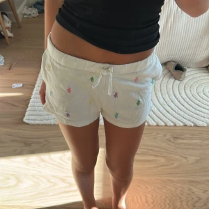Vita shorts med färgglada broderier - Säljer ett par vita shorts med snörning i midjan och små broderade figurer i olika färger som lila, grön, gul och rosa. Modellen är loose fit med slits på sidorna och tillverkad i mjuk bomull, perfekta för chill dagar under sommaren.