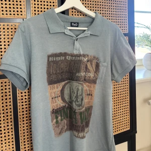 Grå D&G t-shirt med tryck och krage - Snygg grå t-shirt från D&G med coolt vintageinspirerat tryck framtill. Modellen har klassisk pikékrage, knappslå och en liten bröstficka. Perfekt för dig som gillar streetstyle och vill sticka ut med en unik look.