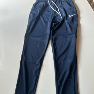 Nike Dri-Fit träningsbyxor marinblå - Marinblå träningsbyxor från Nike med Dri-Fit-teknologi. Byxorna har elastisk midja med vit dragsko och en smal passform. Perfekta för träning eller chill, med klassisk Nike-logga på låret.