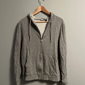 Grå hoodie med dragkedja och huva - Säljer en enkel grå hoodie med dragkedja framtill och två fickor. Tröjan har huva med snörning och är långärmad. Materialet känns mjukt och är troligtvis bomull eller polyester. Perfekt för chill dagar eller när du vill ha något bekvämt på dig.