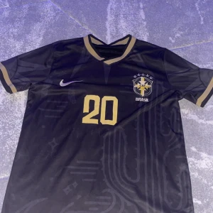 Svart Brasil Vini Jr Nike fotbollströja - Snygg svart Brasilien fotbollströja från Nike med guldiga detaljer och tryck. Nummer 20 och VINI JR på ryggen, samt CBF-emblem på bröstet. Kortärmad med V-ringad krage och guldiga muddar. Tillverkad i lätt, ventilerande polyester som passar perfekt på planen.