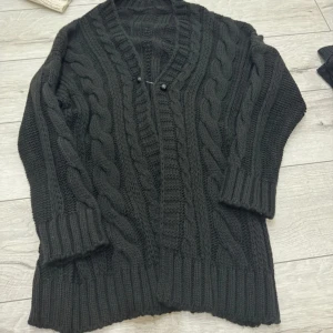 Svart kabelstickad kofta - Mysig svart kabelstickad kofta med grova flätmönster och breda ribbade muddar. Koftan har en djup v-ringning och två små knappar framtill. Perfekt för att hålla värmen under kyliga dagar.