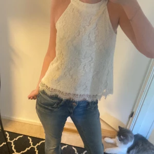 Vit spetsig blus - Supersöt vit blus i spets med hög hals och ärmlös design. Blusen har ett blommigt spetsmönster och avslutas med en fin, lätt fransig kant nertill. Perfekt för dig som gillar romantisk och feminin stil.