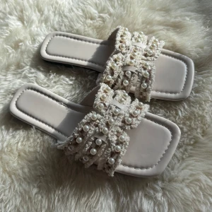 Beige sandaler med pärldetaljer - Superfina beige sandaler med fyrkantig tå och platt sula. Remmarna är dekorerade med pärlor och har en lyxig, texturerad look. Perfekta för att lyfta din sommarstil med en touch av elegans och trendig vibe.