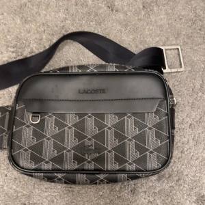 Svart axelväska från Lacoste - Snygg svart axelväska från Lacoste med geometriskt mönster och silverfärgade detaljer. Väskan har justerbart band och dragkedja upptill. Perfekt för att bära det viktigaste och ge en sportig touch till din outfit.