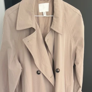 Beige trenchcoat  - Stilren beige trenchcoat med dubbelknäppning och bälte i midjan. Jackan har klassiska detaljer som knappar på ärmsluten och sidofickor. Perfekt för dig som vill ha en tidlös och snygg look under vår och höst.