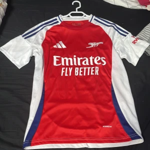 Arsenal fotbollströja Adidas S - Säljer en Arsenal matchtröja från Adidas i slim fit. Tröjan är röd framtill och baktill med vita och blå detaljer på ärmarna. Klassisk rund hals och korta ärmar. Materialet är lätt och andas, perfekt för träning eller match. Emirates Fly Better tryck på bröstet.