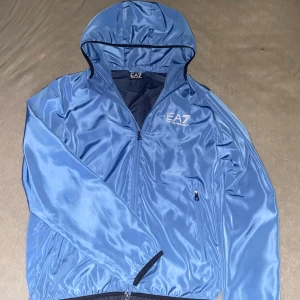 Blå vindjacka från EA7 Emporio Armani - Snygg blå vindjacka från EA7 Emporio Armani med huva och dragkedja framtill. Jackan har logga på bröstet, meshfoder och två fickor med dragkedja. Perfekt för dig som vill ha en stilren och sportig look.