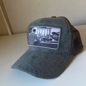 Grå velvetkeps med foto-patch - Snygg grå keps med mjuk mockaliknande front och mesh på sidorna och bak. Framtill sitter en patch med svartvitt foto av en person i randig tröja framför klassiska pelare. Perfekt för dig som gillar unik streetstyle. Det är dessvärre vit färg på insidan av kepsen som inte syns.