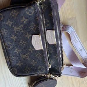 Louis Vuitton monogram axelväska - Säljer en brun axelväska från Louis Vuitton med klassiskt monogrammönster och guldiga detaljer. Väskan har flera fack med dragkedja och en bred, ljusrosa axelrem med logga. Materialet är skinn och syntet, perfekt för dig som vill ha en ikonisk och trendig accessoar.