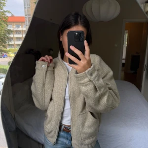 Beige teddyjacka med dragkedja - Mysig beige teddyjacka med dragkedja framtill och stora fickor. Jackan har en avslappnad passform och ribbade muddar vid ärmslut och midja. Perfekt för lager-på-lager och ger en chill vibe till din outfit.