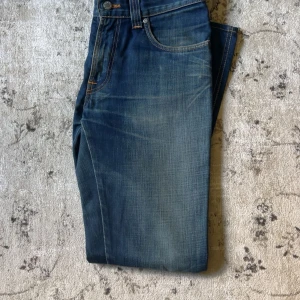 Blåa nudie jeans - Säljer ett par klassiska blå jeans från Nudie Jeans i storlek 31/32. Jeansen har en snygg tvätt med lätt slitning och orangea kontrastsömmar. Modellen har normal passform och raka ben, perfekt till sneakers eller boots.
