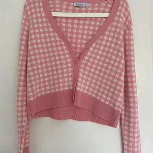 Rutig rosa kofta från Stradivarius - Superfin croppad kofta från Stradivarius i rosa och vitt rutigt mönster. Koftan har v-ringad hals, långa ärmar och knappar framtill. Mjuk stickad kvalitet som känns skön mot huden och passar perfekt till jeans eller kjol.