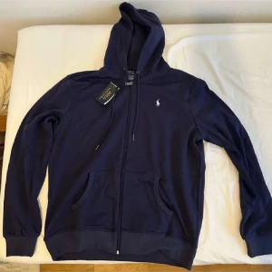 Marinblå hoodie från Polo Ralph Lauren - Säljer en marinblå hoodie från Polo Ralph Lauren med klassisk logga på bröstet. Tröjan har huva med snörning, dragkedja framtill och två fickor. Tillverkad i mjuk bomull, perfekt för chill dagar. Långärmad och stilren design.