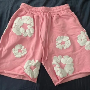 Rosa shorts från Denim Tears med blommor strl S - Säljer ett par rosa shorts från Denim Tears med vita blommiga applikationer över hela plagget. De har elastisk midja med snörning och är tillverkade i mjuk bomull. Perfekta för sommarens chill och har en loose passform som ger skön vibe.