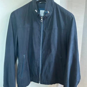 Svart vindjacka från Bugatti, strl 54 - Snygg svart vindjacka från Bugatti i storlek 54. Jackan har dragkedja framtill, hög krage med snörning och två fickor med dragkedja. Tillverkad i 100% polyester, perfekt för blåsiga dagar. Enkel och stilren design som funkar till det mesta. nyskick