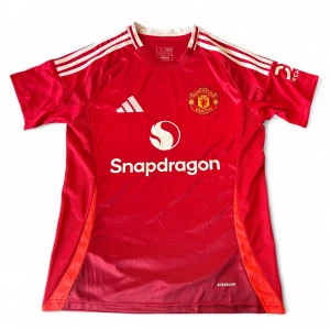 Manchester United röd matchtröja Adidas - Snygg Manchester United fotbollströja från Adidas i röd färg med vita detaljer och klubbmärke på bröstet. Kortärmad med klassiska tre ränder på axlarna och Snapdragon-logga framtill. Tillverkad i lätt och ventilerande polyester, perfekt för träning eller match.