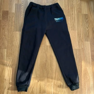 Svarta mjukisbyxor Trapstar - Svarta mjukisbyxor från Trapstar med blå och vit logga på ena benet. Byxorna har resår i midjan och vid bensluten, samt sidofickor. Sköna och avslappnade, perfekta för chill eller streetwear. Materialet är mjukt och känns riktigt bekvämt.