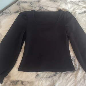 Svart långärmad topp med puffärm - Stilren svart topp med långa puffärmar och fyrkantig ringning. Snygg passform som markerar midjan och ger en clean look. Perfekt att styla med jeans eller kjol för en trendig outfit. Toppen har aldrig kommit till användning!