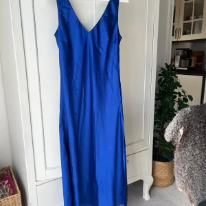 Blå långklänning med v-ringning - Snygg blå långklänning med djup v-ringning och breda axelband. Klänningen är ärmlös och har en rak siluett som faller längs kroppen. Materialet har ett lätt glansigt tyg. Aldrig använd!🥰🥰
