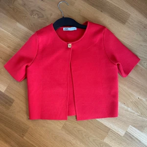 Röd bolerotopp från Zara - Kort röd bolerotopp från Zara med rund halsringning och en dekorativ guldig knapp framtill. Tjockare material och korta ärmar ger en stilren och modern look. Perfekt för att lyfta en outfit med en färgklick.