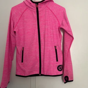 Rosa vindjacka med huva  - Snygg rosa vindjacka med svart dragkedja och detaljer. Jackan har huva, två fickor med dragkedja och tumhål vid ärmslut. Perfekt för dig som gillar sportig stil. 