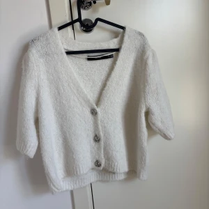 Vit kofta med kort ärm - Supermjuk och fluffig vit kofta från vero Moda med tre snygga knappar framtill. Koftan har v-ringning, korta ärmar och en croppad passform som ger en chill vibe. Perfekt att slänga över ett linne eller t-shirt för en enkel men stilren look.